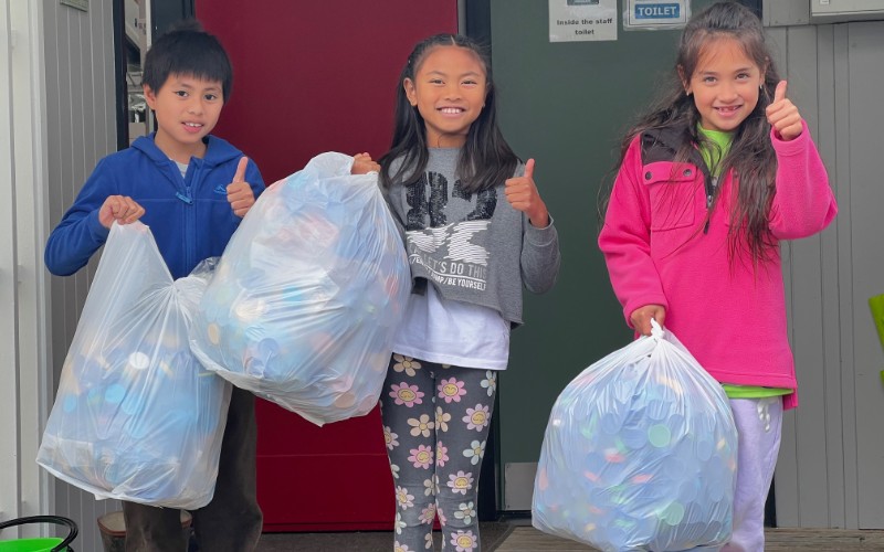 Three kids holding white plastic bags full of milk bottle lids.